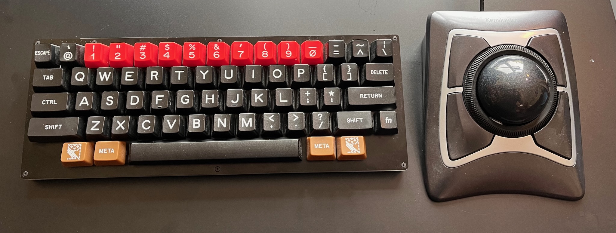 a dusty compact keyboard with red number keys and brown modifier keys; the ALT keys feature the BBC computer literacy project owl logo; to the right of the keyboard is a Kensington Expert Mouse trackball