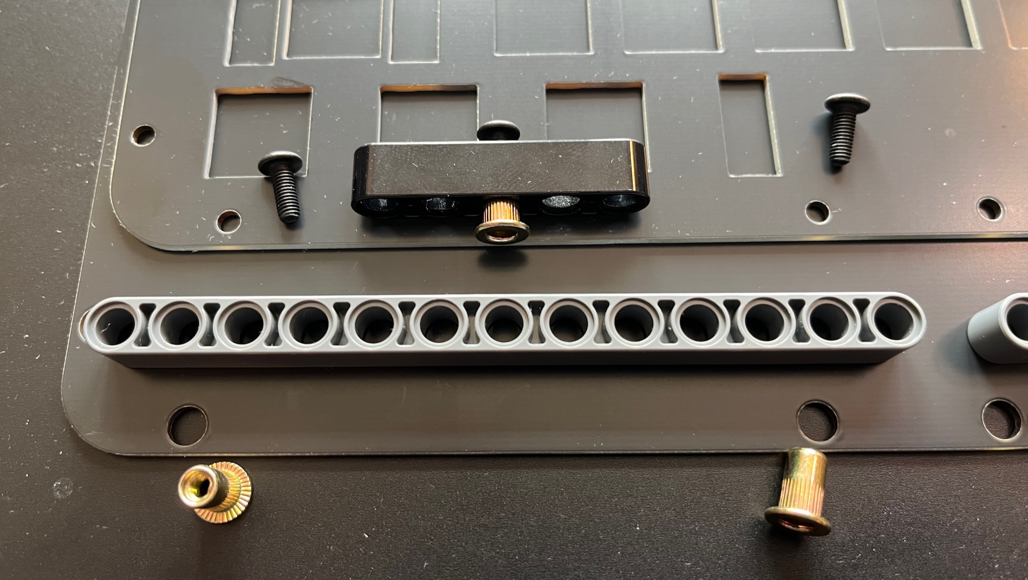 un-assembled parts of keybird69: a black switch mounting plate on top of a black base plate, with a lego beam and some screws and rivet nuts placed to show how they will fit together