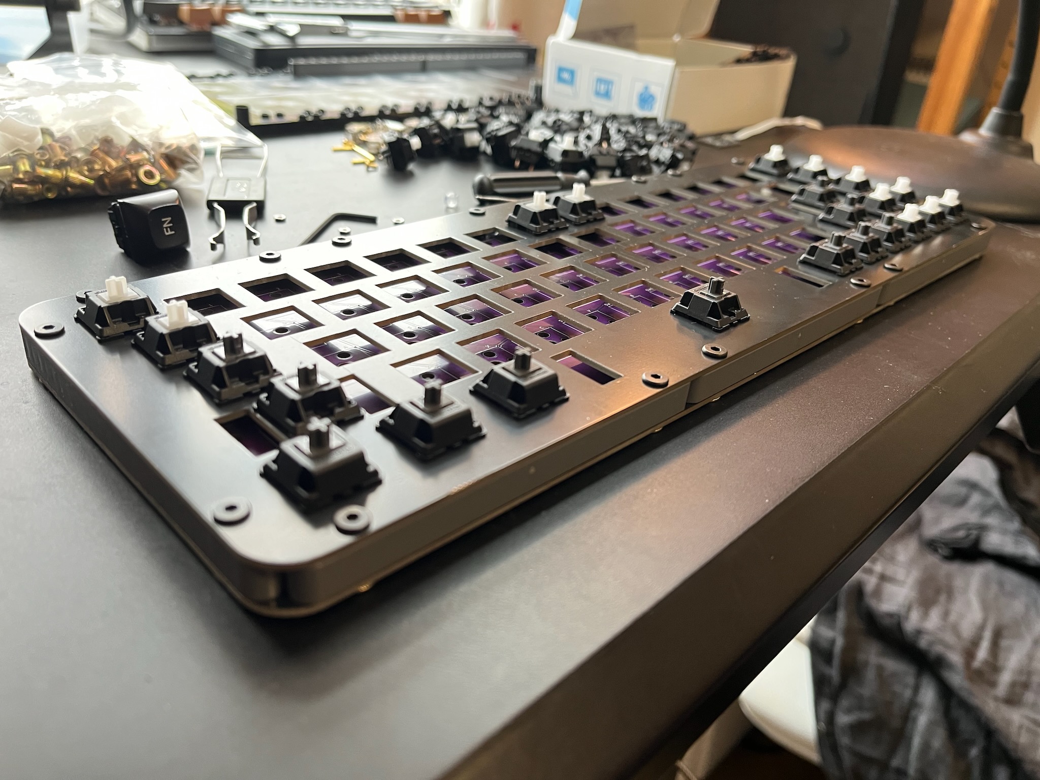a partially-assembled keybird69; the case is screwed together and a couple of dozen switches are in place; the purple PCB is visible through the unoccupied holes in the switch mounting plate; behind the keyboard is a clutter of switches, tools, fasteners, lego ...