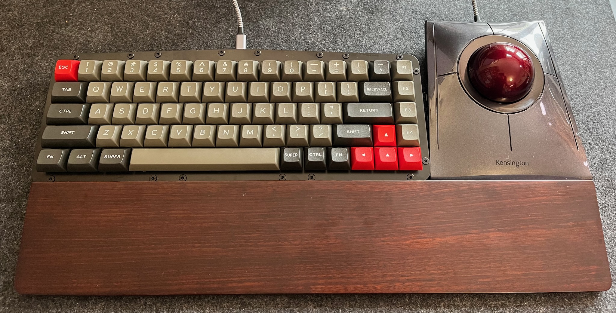 a compact keyboard; most of the keycaps are warm grey with white legends, but the modifiers and special keys are black and white, escept for escape and arrows which are red and white; to the right is a Kensington Slimblade trackball, and below both of them is a dark wooden wrist rest